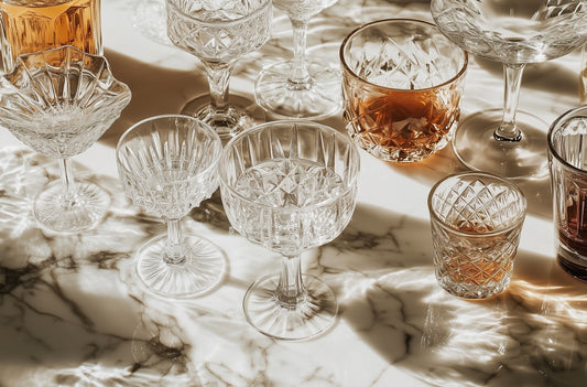 A variety of crystal glassware on a marble surface, including coupe glasses, tumblers, and highballs, illustrating the elegance and function of different drink-specific glasses.